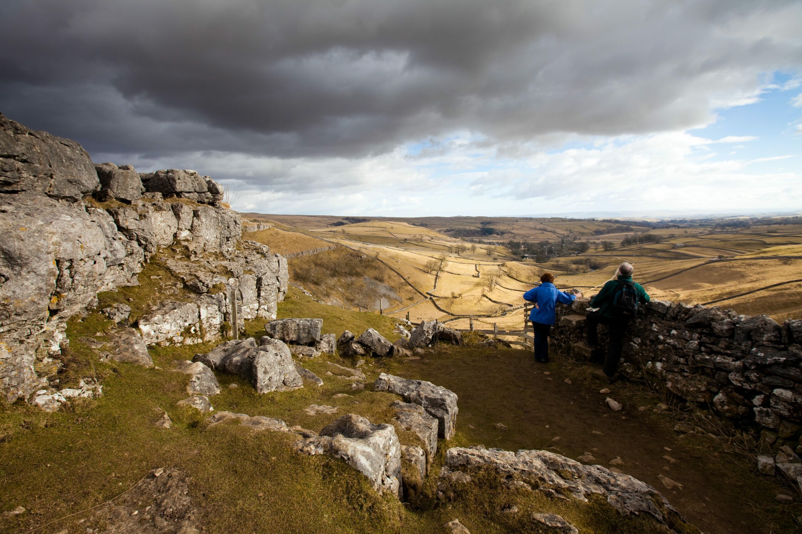 Two hikers looking at a panoramic view from atop a rocky hill under cloudy skies.