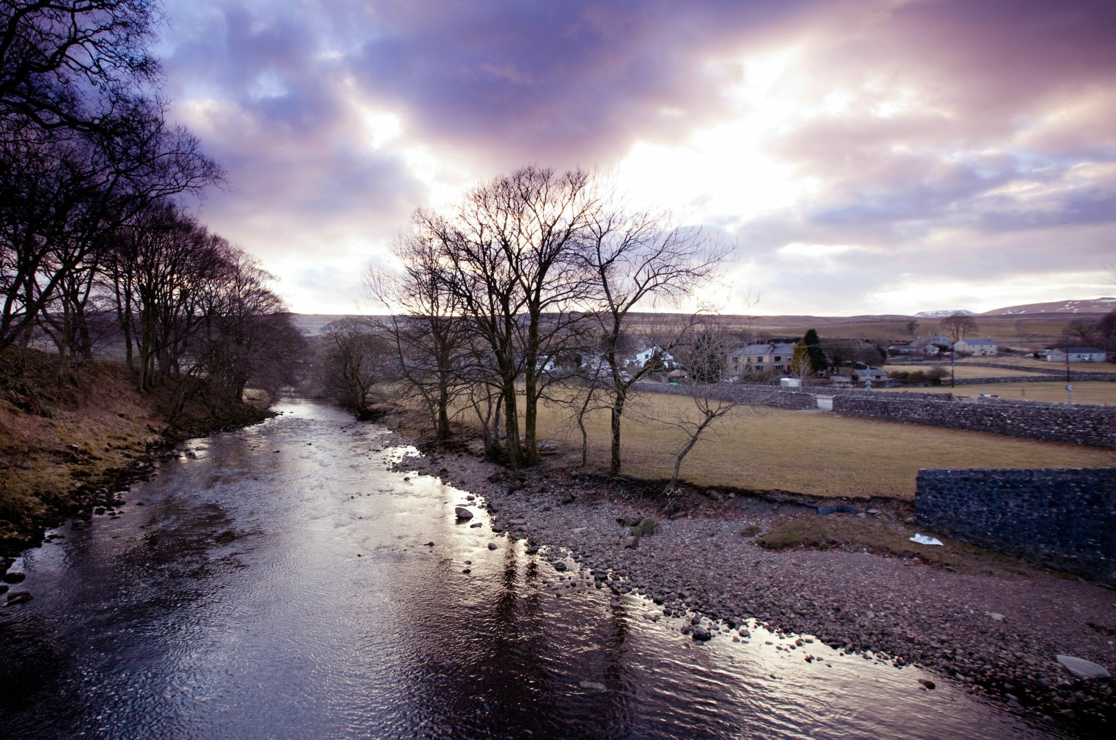 Sunset over a serene river with bare trees and a small village in the background.