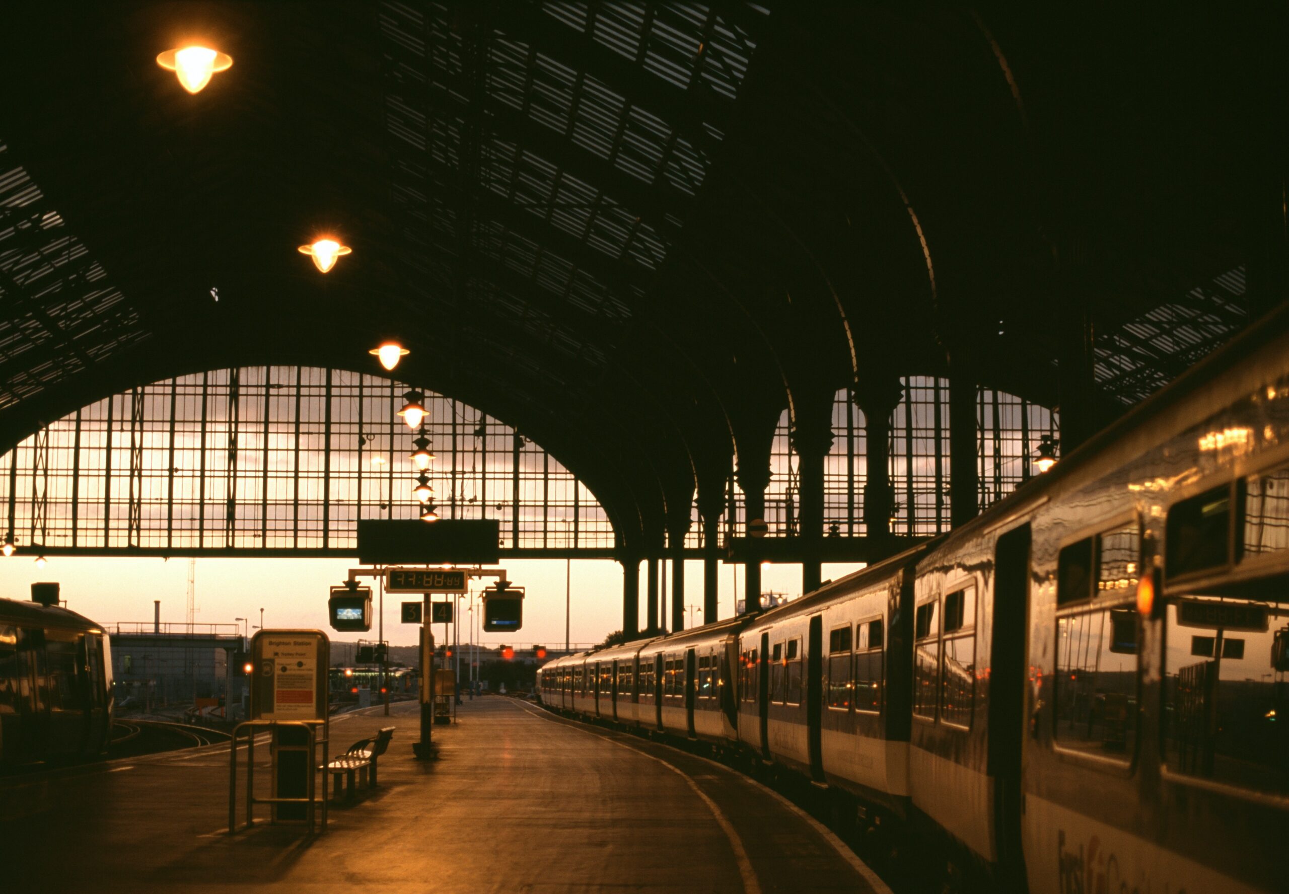 A train is stationary on a platform inside a large, arched train station at dusk. The station is dimly lit with overhead lights and the sky is visible through a glass facade.